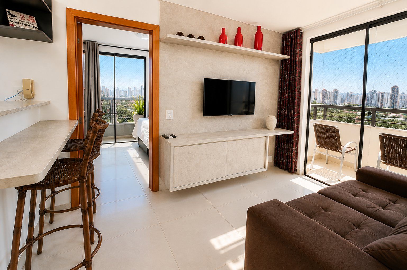 A modern living room with a flat-screen TV, a dining area with wicker chairs, a sofa, and a balcony with a city view.