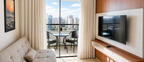 A modern living room with a sofa, a flat-screen TV, a dining table with chairs, and a balcony with a city view.