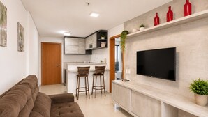 Un salon moderne avec un canapé, un téléviseur fixé sur un meuble et un coin cuisine équipé de tabourets.