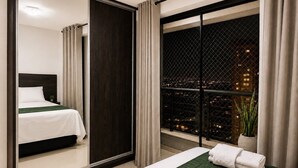 Une chambre d’hôtel avec un lit, un grand miroir, un balcon offrant une vue sur la ville la nuit, et une plante en pot.