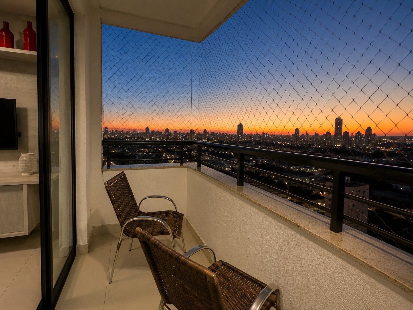 A balcony with wicker chairs, a glass door, and a cityscape view at sunset.