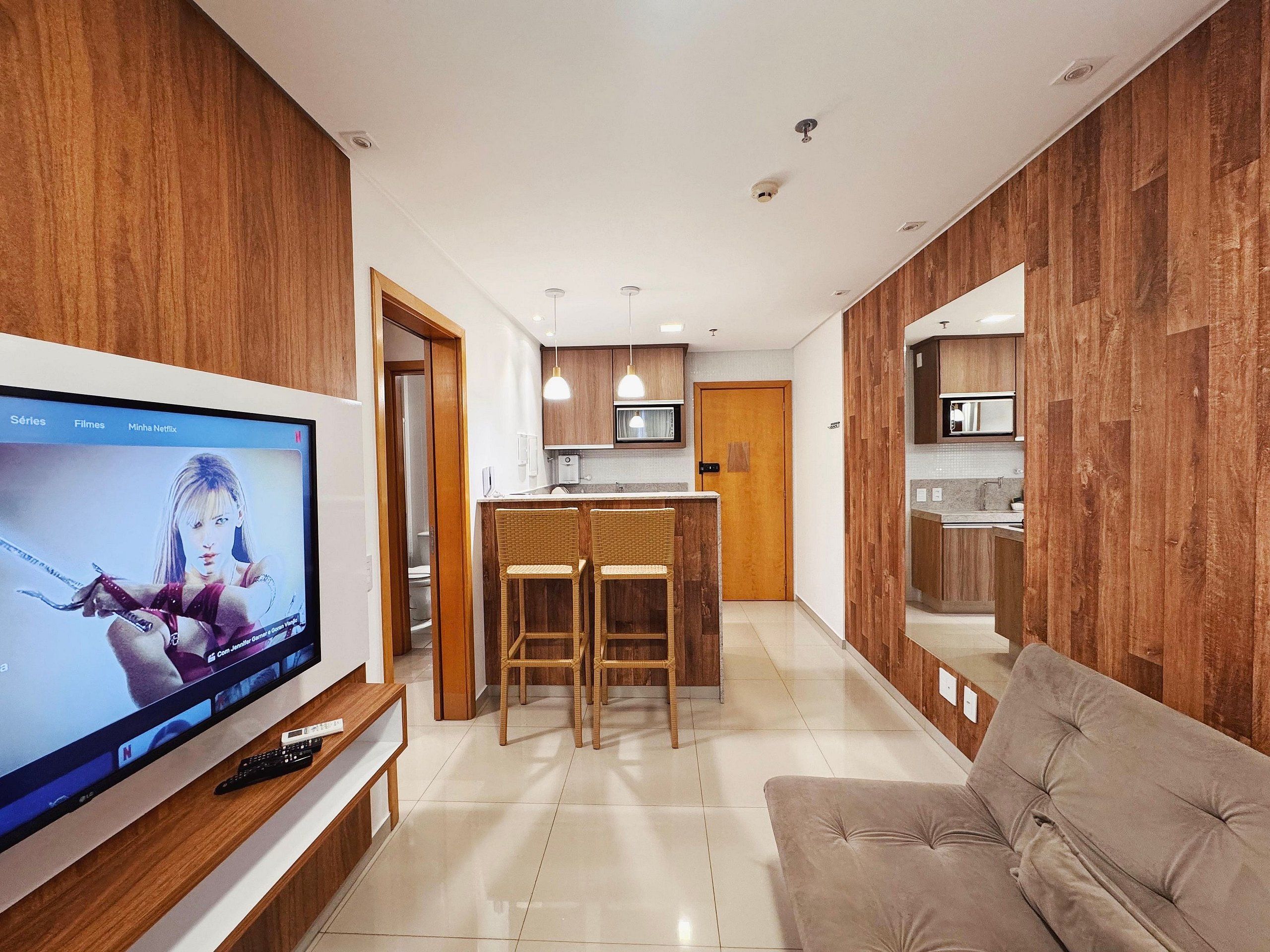 Uma sala de estar moderna com televisão de tela plana, sofá, área de cozinha com armários de madeira e espaço de jantar com duas cadeiras.