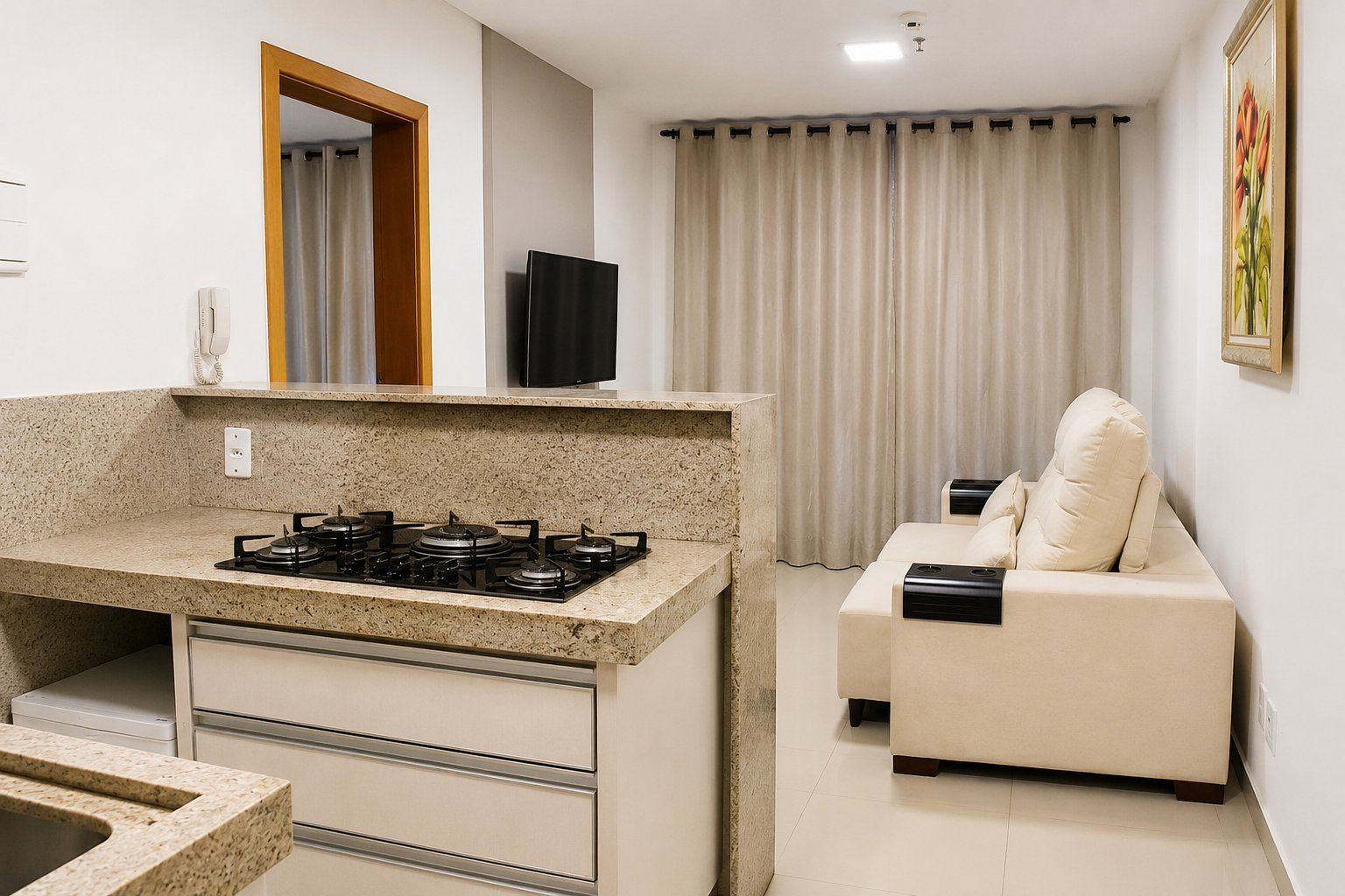 A compact kitchen with a gas stove, a small countertop, and a built-in oven. There is a beige sofa with a cushion and a flat-screen TV mounted on the wall.