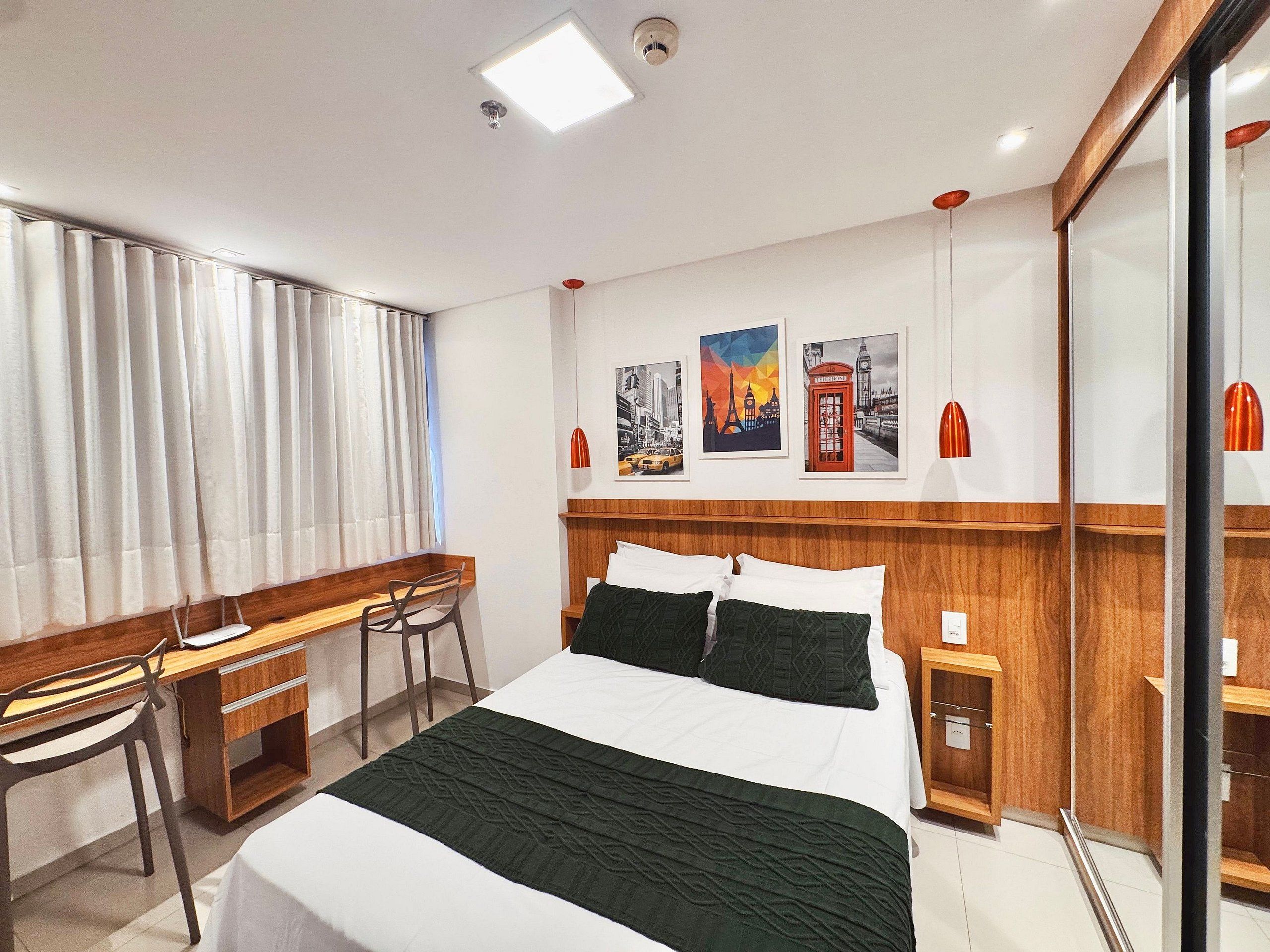 Quarto de hotel com cama, escrivaninha, cadeira e uma parede com quadros.