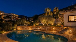 Piscina com paredes de pedra e escadas, rodeada por vegetação e um edifício com fachada branca.