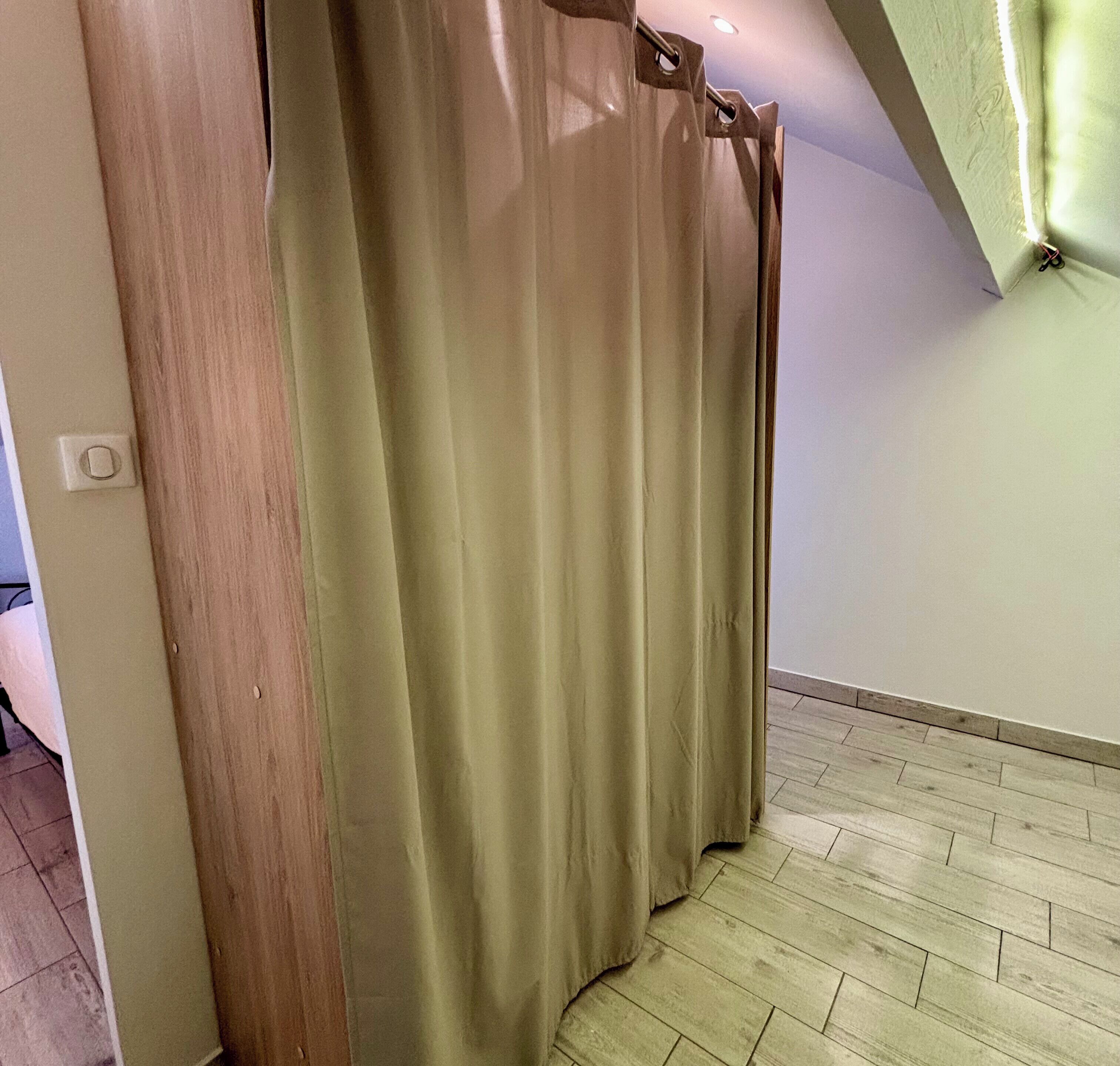 Ein Zimmer mit Holztür, beige farbenen Vorhängen, einem hellen Boden und einer weißen Wand.