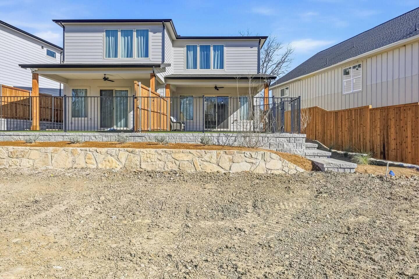 A two-story residential building with a stone retaining wall, a wooden fence, and a clear sky.