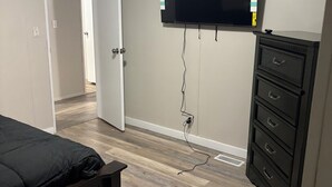 Ein Schlafzimmer mit einem Bett, einem Schminktisch, einem an der Wand montierten Fernseher und einer Tür zu einem anderen Raum.