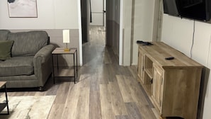 Ein Wohnzimmer mit grauem Sofa, einem kleinen Tisch mit Lampe, einem Holzschrank und einem an der Wand befestigten Fernseher.