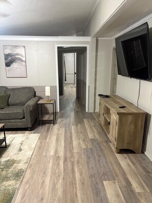 A living room with a grey sofa, a small table with a lamp, a wooden cabinet, and a TV mounted on the wall. -  3-bedroom 2 full bath mobile home in nice quiet area 10 minutes from town (Richwood)