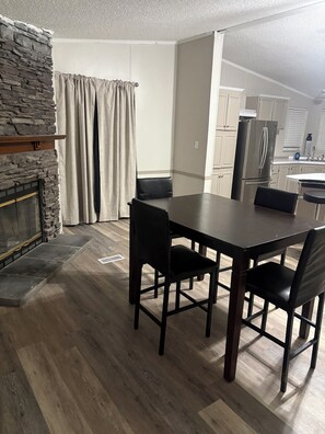 A dining area with a stone fireplace, a table with chairs, and a kitchen in the background. -  3-bedroom 2 full bath mobile home in nice quiet area 10 minutes from town (Richwood)