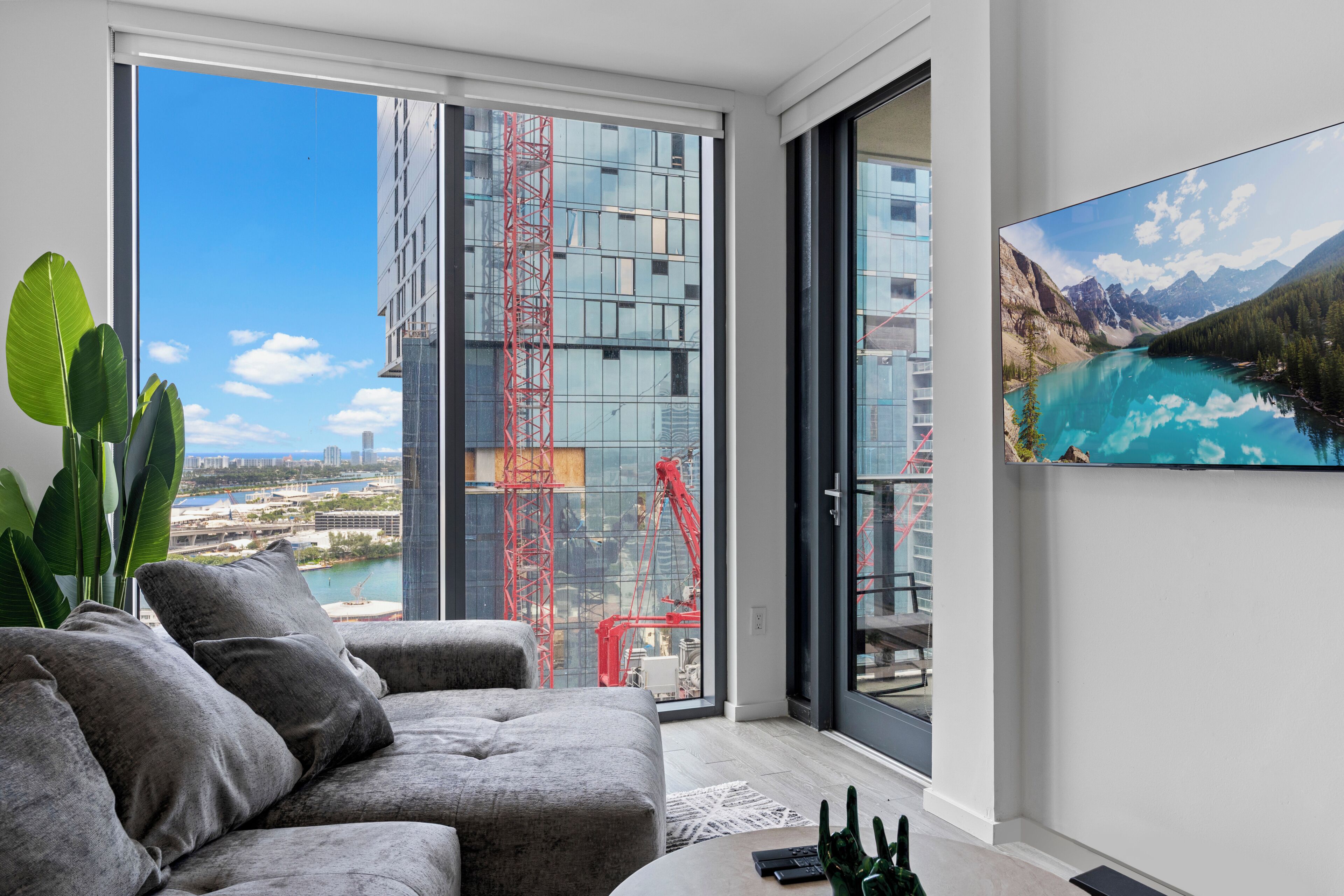Ein modernes Wohnzimmer mit einem grauen Sofa, einem Couchtisch und einem großen Fenster mit Blick auf eine Baustelle.
