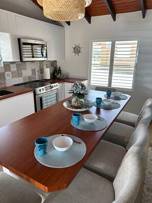 A modern kitchen with white cabinets, a built-in oven, and a dining area with a long wooden table and grey upholstered chairs.