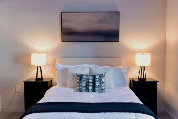 A neatly made bed with white linens, a dark blue throw blanket, and a patterned pillow. There are two bedside tables with lamps, a black-framed painting above the bed, and a plain wall.