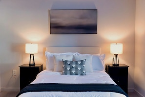 Un lit bien fait, avec du linge de lit blanc, un plaid bleu foncé et un coussin à motifs. On y trouve deux tables de chevet avec des lampes, un tableau encadré de noir au-dessus du lit et un mur sobre.