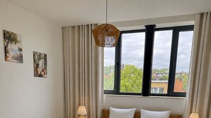 Un dormitorio con una cama grande, dos mesitas de noche, una lámpara colgante y una ventana con vistas a edificios y árboles.