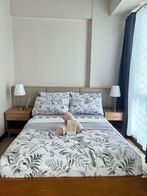 A neatly made bed with floral bedding, two bedside lamps, and a nightstand with a lamp and a box. - Mactan Infinity Pool View|Near Airport & Beach (Lapu-Lapu)