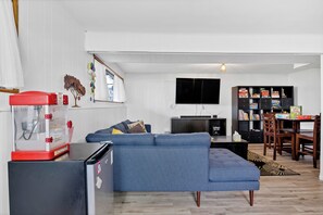 Ein Wohnzimmer mit blauem Sofa, Flachbildfernseher, Bücherregal und einem Essbereich mit Holztischen und Stühlen.