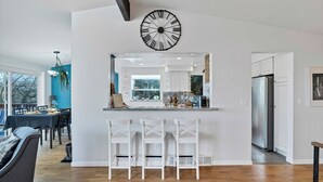 A modern kitchen with a large clock, white bar stools, and a dining area with a table and chairs. - Seattle Skyline & Lake Retreat | 3Bd/3Ba (Seattle)