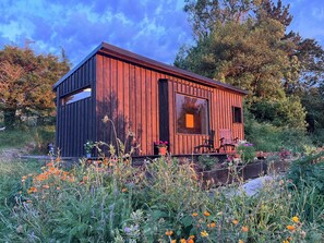 Een houten blokhut met een groot raam, omgeven door een tuin vol verschillende bloemen en planten.
