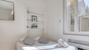 Un dormitorio ordenado con cama, almohadas, una estantería con objetos decorativos, una ventana y un espejo adosado a la pared.