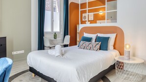 Ein modernes Hotelzimmer mit einem großen Bett, einem kleinen Tisch und einem Stuhl, einem Fenster mit Vorhängen und einem an der Wand befestigten Regal mit Dekorationen.