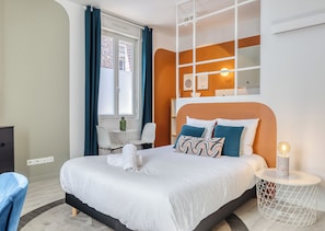 Une chambre d’hôtel moderne avec un grand lit, une petite table et une chaise, une fenêtre avec des rideaux et une étagère fixée au mur, ornée d’objets décoratifs.