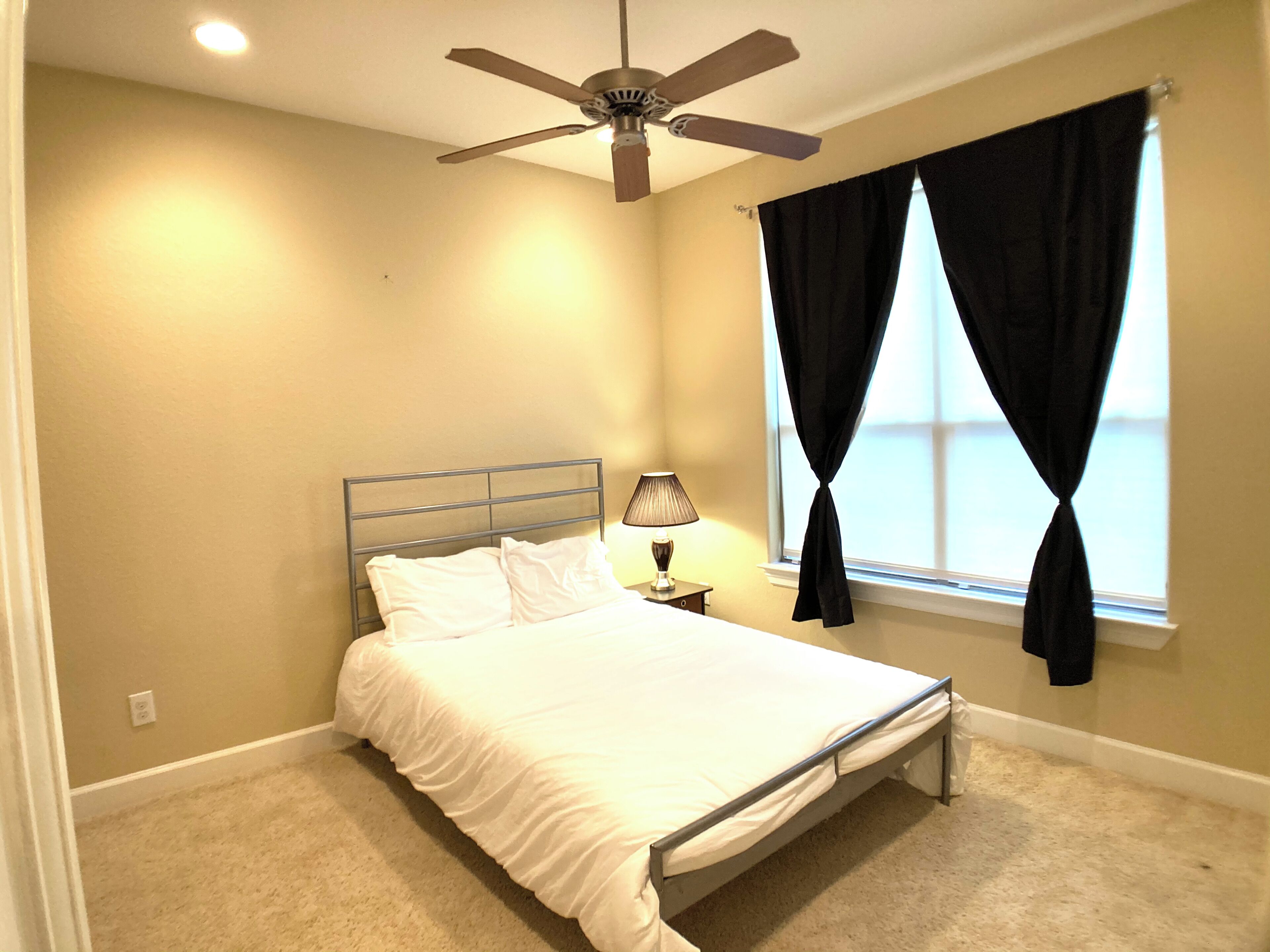 A bedroom with a bed, a nightstand, a lamp, a ceiling fan, and a window with curtains.