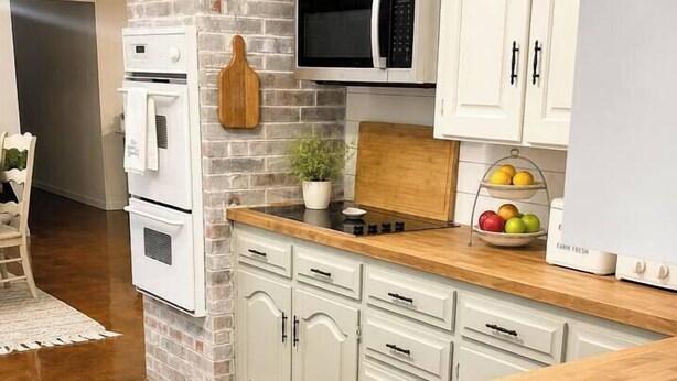 A kitchen with white cabinets, a brick accent wall, a microwave, and a fruit bowl. - Shady Pecan Farmside (Red Oak)