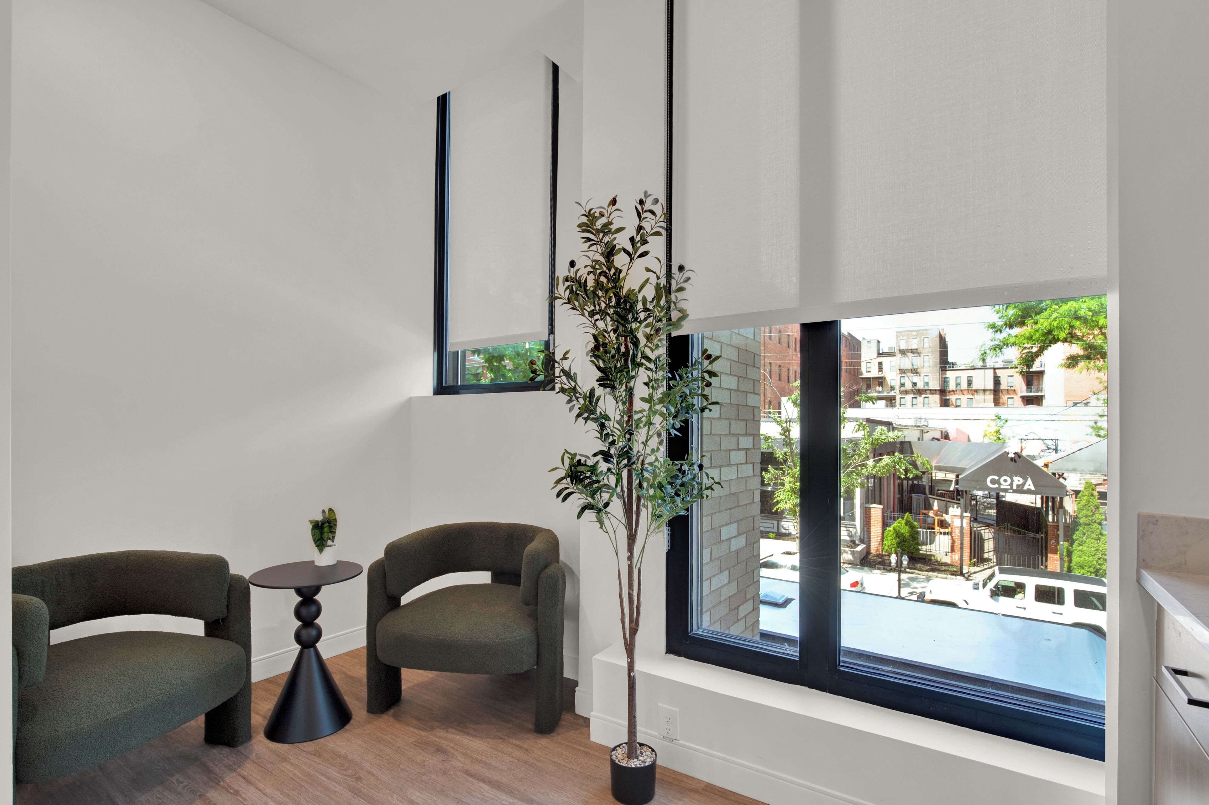 A modern room with two green armchairs, a small round table, a potted plant, and a large window offering a view of the city.