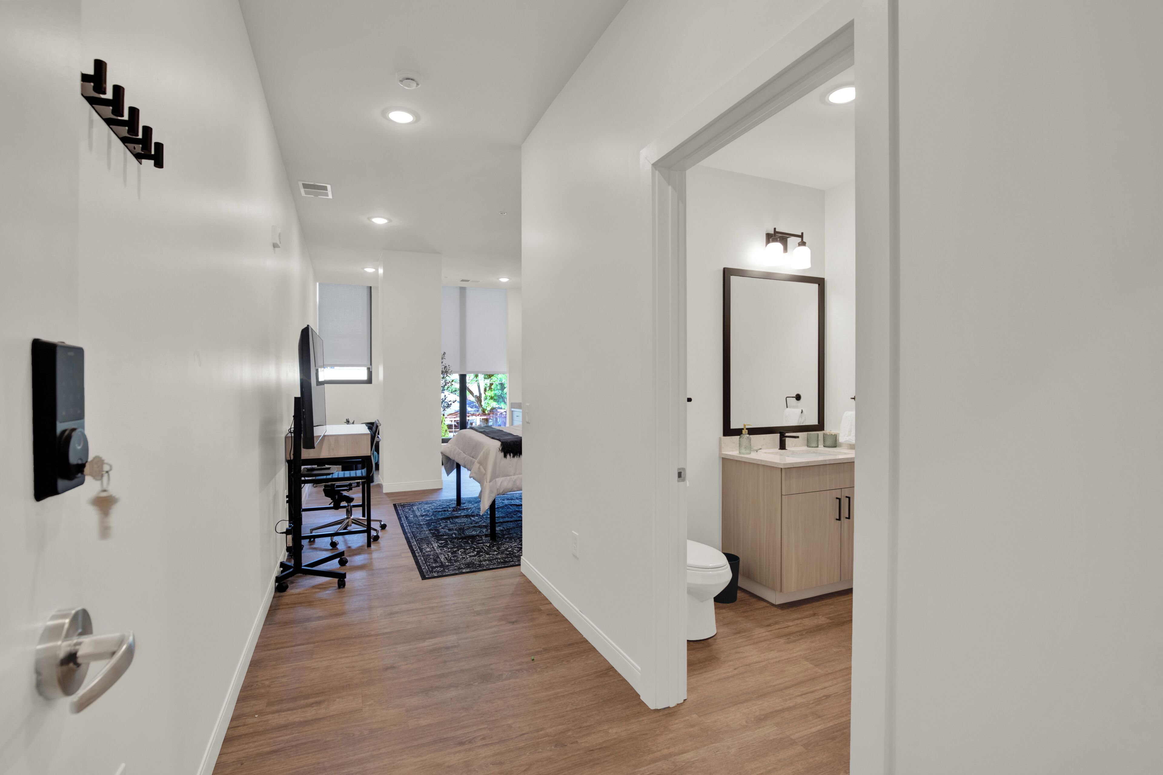 A modern hotel room with a wooden floor, a desk with a chair, a bed, and a bathroom with a sink and mirror.