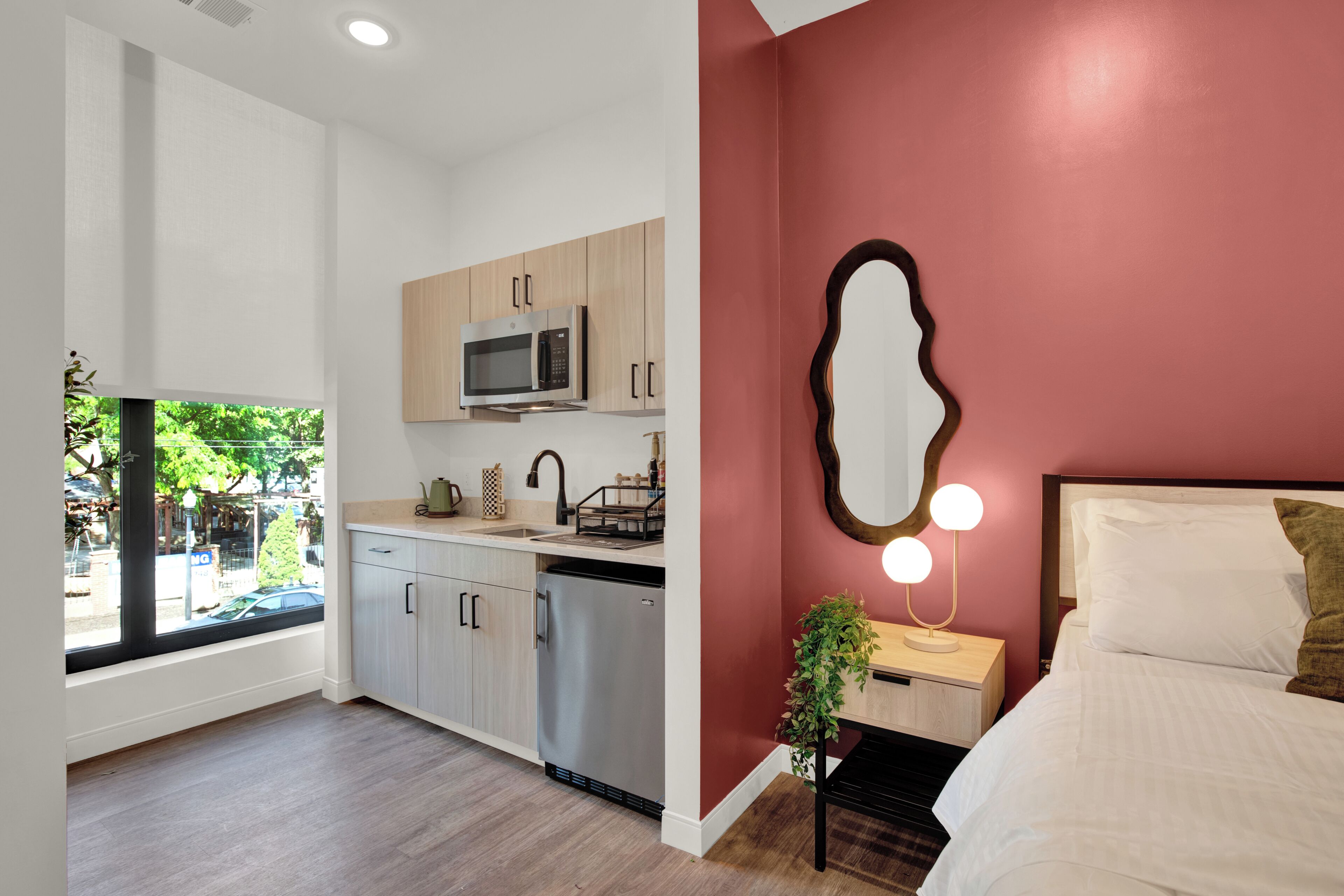 A modern room with a kitchen, a large window, a bed, and a unique wall mirror.