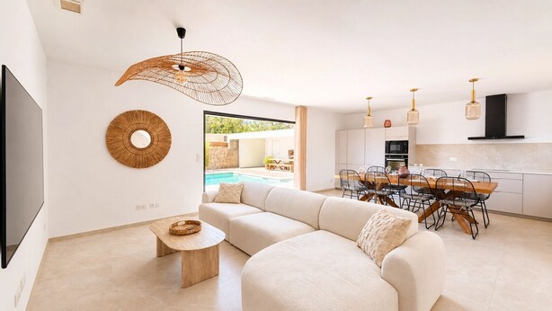 Ein modernes Wohnzimmer mit einer Eckcouch, einem Essbereich mit Holztisch und -stühlen, einer Küche mit schwarzem Dunstabzug und einem großen Fenster mit Blick auf einen Pool.