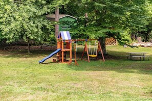A playground with a slide and swings, a picnic table, and a green house in the background.