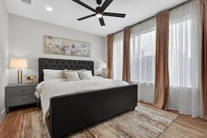 Ein Schlafzimmer mit Bett, Nachttischen, Deckenventilator, Gemälde und durchsichtigen Vorhängen.