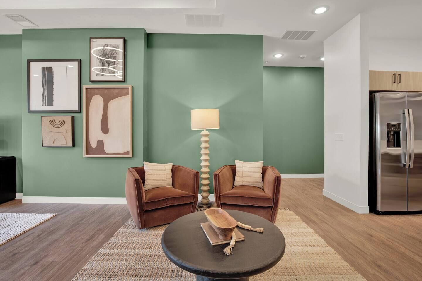 A modern living room with two brown armchairs, a round coffee table, and a floor lamp. A large green wall with framed artwork is behind the seating.