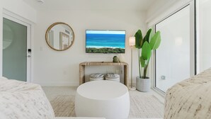 A modern living room with a round white ottoman, a flat-screen TV on the wall, a console table with decorative items, a large potted plant, and a sliding glass door. - The Arches: Waterfront Suite in Boca (Boca Raton)