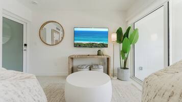A modern living room with a round white ottoman, a flat-screen TV on the wall, a console table with decorative items, a large potted plant, and a sliding glass door.
