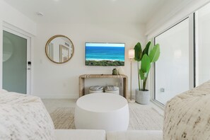 Ein modernes Wohnzimmer mit einem runden weißen Hocker, einem Flachbildfernseher an der Wand, einem Konsolentisch mit Dekoration, einer großen Topfpflanze und einer Glastür.