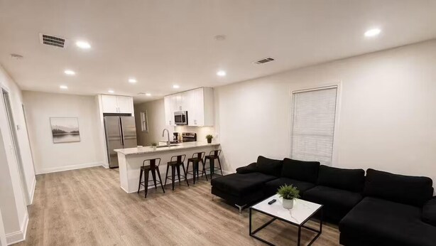 Una sala de estar moderna con un sofá negro, una mesa de centro y una zona de cocina con refrigerador, microondas y taburetes de bar.