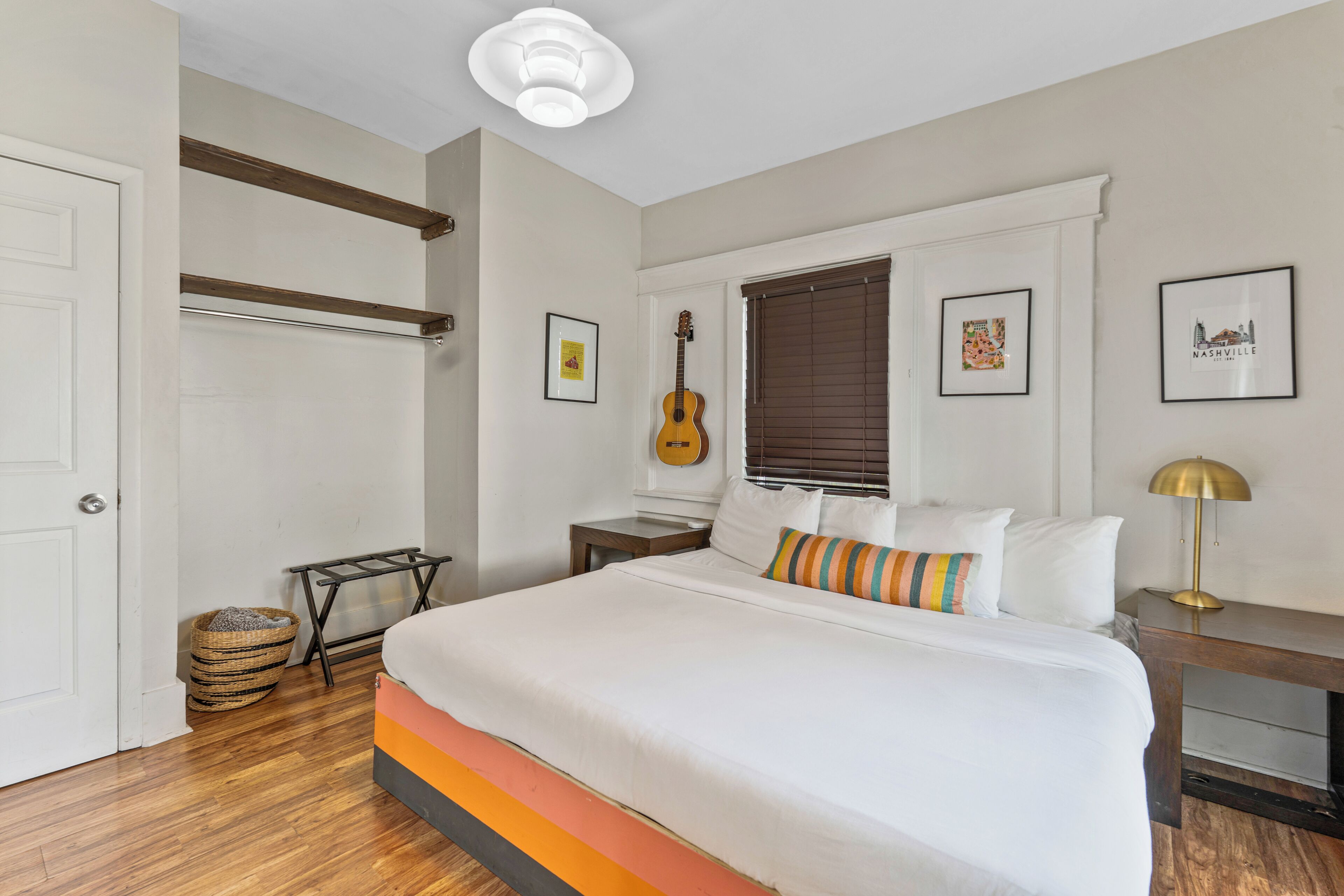 Een slaapkamer met een bed, een gitaar, een raam met jaloezieën, ingelijste foto's en een lamp.