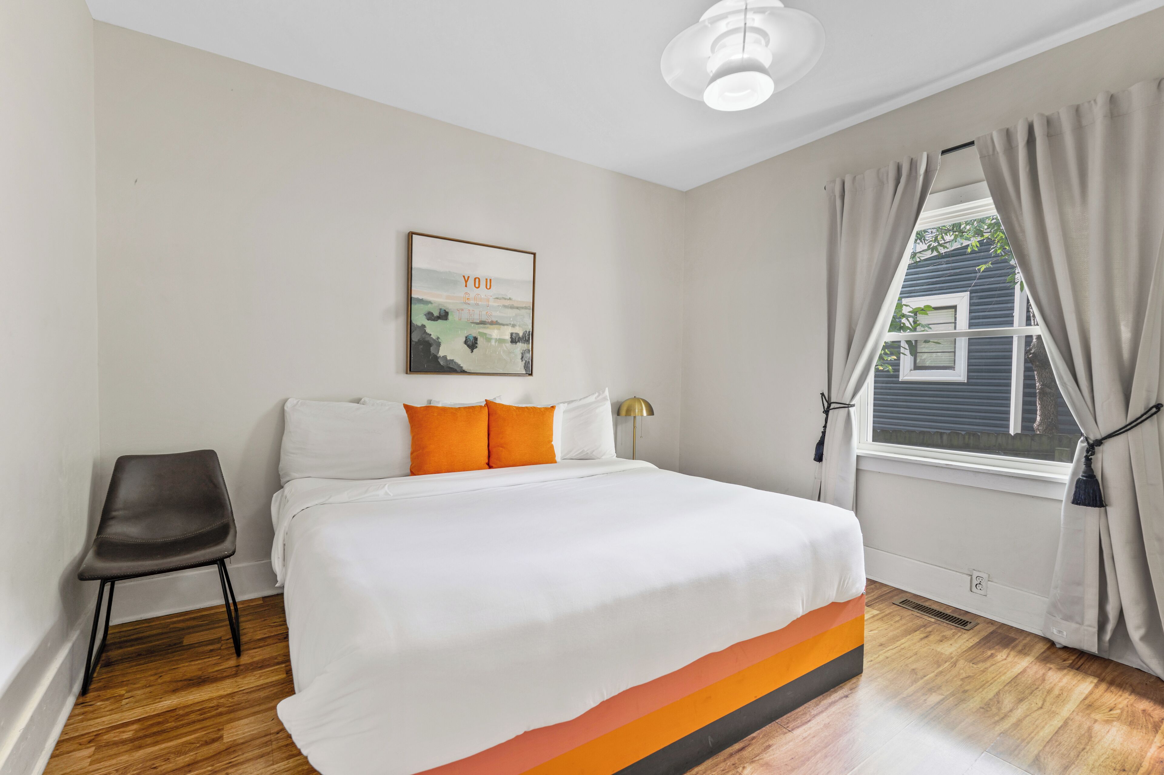 A bedroom with a bed, a chair, a window with curtains, a wall-mounted light fixture, and a framed picture.