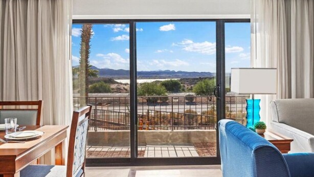A room with a dining table, chairs, a sofa, a lamp, and a view of a landscape through large windows.