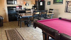 A pool table with a red felt surface, a ceiling fan, wooden cabinets, a kitchen area with appliances, and a dining table with chairs. - Northern Lights Lodge (Watford City)