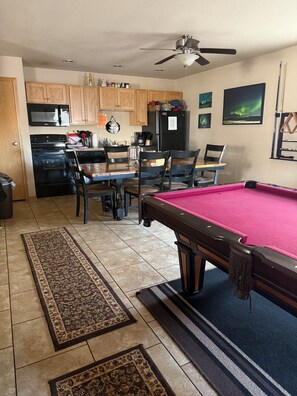 A pool table with a red felt surface, a ceiling fan, wooden cabinets, a kitchen area with appliances, and a dining table with chairs. - Northern Lights Lodge (Watford City)