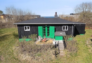 Ein kleines Holzgebäude mit schwarzem Dach und grünen Fensterläden, umgeben von Gras und Sträuchern.