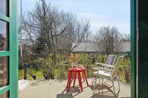 Eine Terrasse mit einem roten Tisch und Stühlen, einem weißen Sessel und Blick auf Bäume sowie ein Gebäude im Hintergrund.