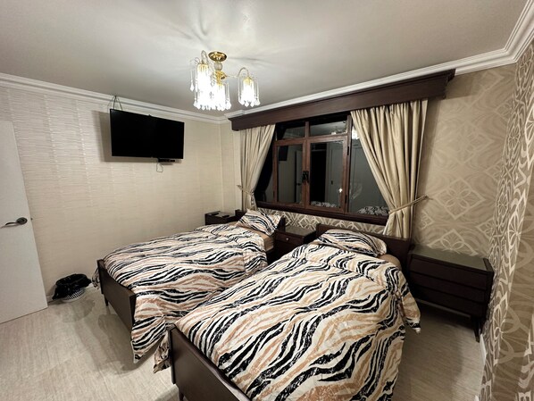 A bedroom with two beds, a TV mounted on the wall, a window with curtains, and a chandelier.