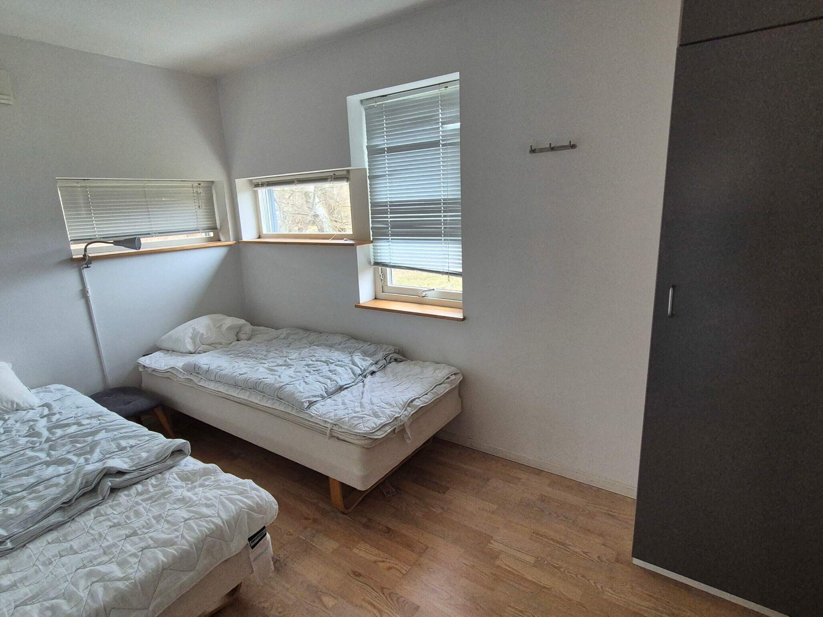 A small, single-bed room with a wooden floor, two windows with blinds, and a dark-colored wardrobe.