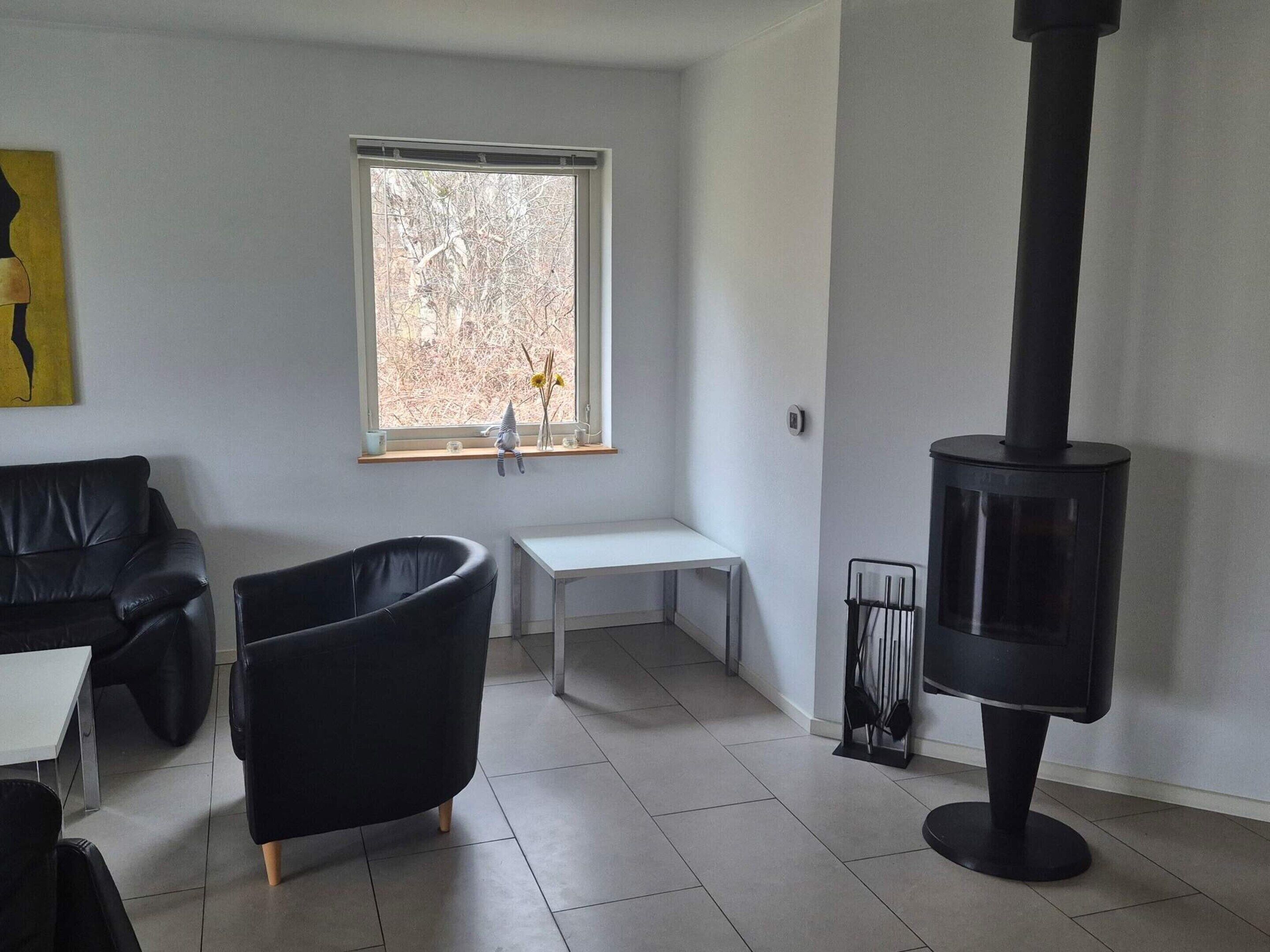 A modern living room with a black sofa, a black armchair, a white table, a window with a view of trees, and a black wood-burning stove.